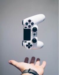 A white gaming controller with black buttons and analog sticks is levitating above a person's open hand against a grey background.